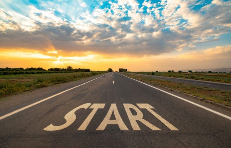 Sunset over a straight road extending to the horizon, with large 'START' letters painted on the pavement in the foreground.